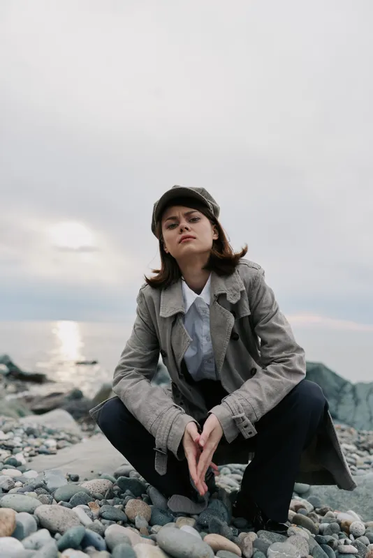 Young woman wears coat and hat sitting on rocky beach looking confident with cloudy sky and calm sea in background. Concept for fashion, outdoor, and autumn style.