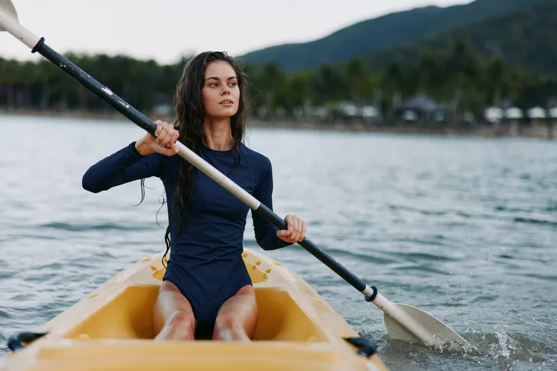 A young woman paddles a yellow kayak on calm water, wearing a navy swimsuit, gripping the paddle with a determined posture as distant hills blur on the horizon.