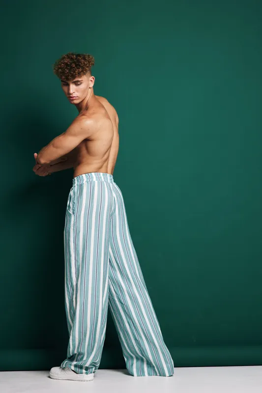 Male model, young and shirtless with curly hair posing in vertical striped wide pants and white sneakers standing against dark green background in studio photo shoot setting.