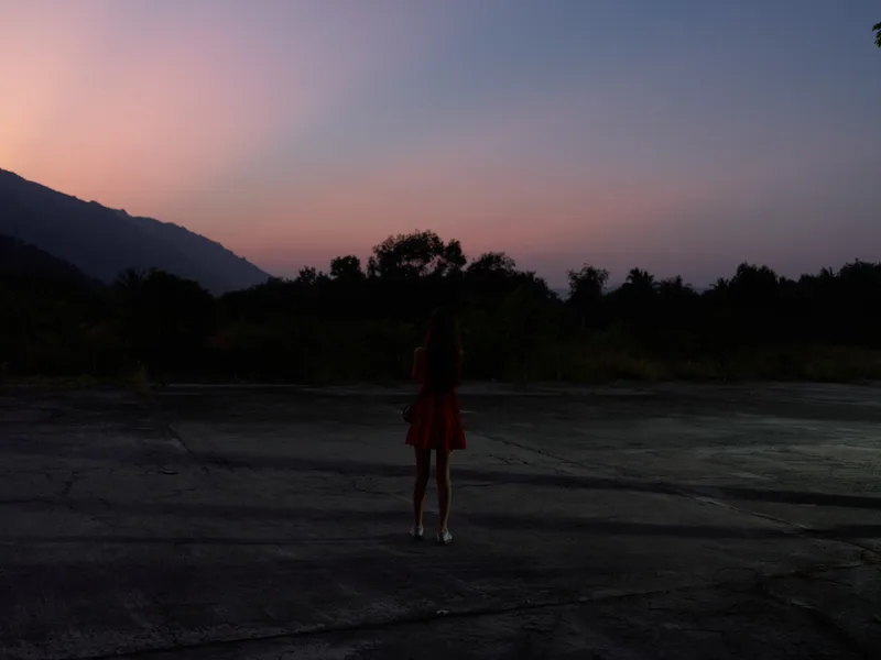 silhouette of a woman standing alone on an empty concrete surface at dusk, with mountains and trees in the background under a colorful twilight sky