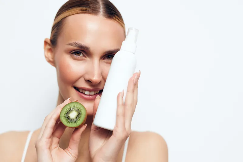 Bright smiling woman holds a kiwi beside a white skincare product bottle, showcasing natural beauty and a fresh glow in a clean studio setting for a modern beauty shoot