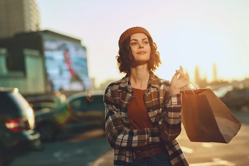 Woman shopping urban candid lifestyle authenticity mindful golden glow young woman with shopping bags smiling in city parking at sunset, relaxed confident portrait with warm emotional storytelling.