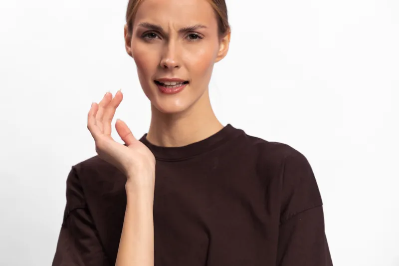 Confused woman in dark shirt looks uncertain and raises hand in questioning gesture. Studio portrait on white background with expression of doubt and hesitation. Concept of confusion and indecision.