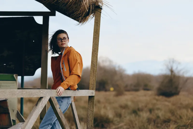 Woman lifestyle outdoor portrait, standing on a wooden deck, orange jacket and jeans, glasses, curly hair, soft calm sunlight and rural countryside background, winter air, bright daytime.