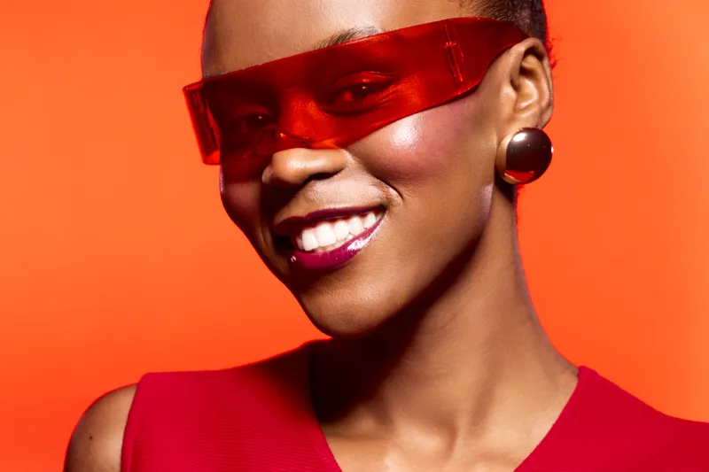 Smiling black woman with dark skin wearing red futuristic sunglasses and red top poses against bright orange background. Concept for fashion, beauty, and modern style with vibrant colors.