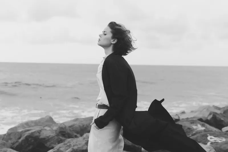 Woman standing on a rocky coast facing the sea, wind in her hair, wearing a flowing coat and dress, monochrome mood with open horizon