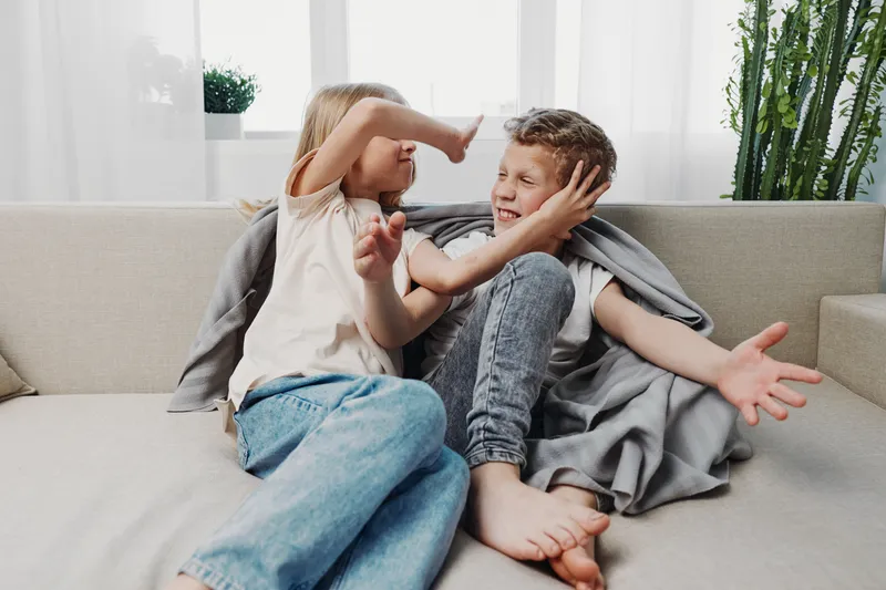 Playful siblings sitting on couch in casual clothes, boy and girl having fun and joking, bright living room with natural light and plants in background.