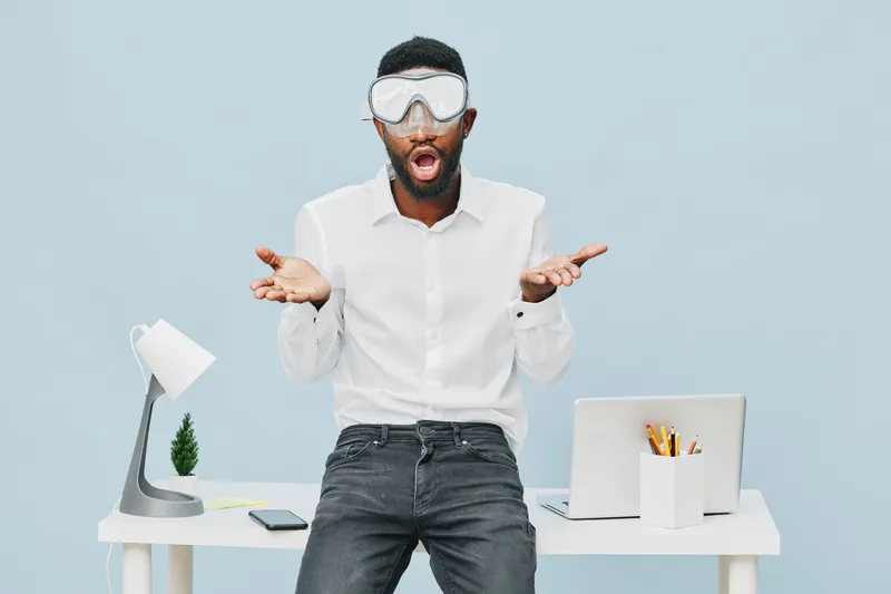 Man wearing white shirt and protective goggles expresses surprise while standing at modern office desk with computer, smartphone, and stationery on light blue background. People, office, technology co