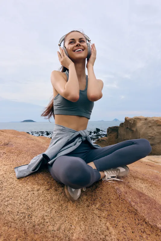 Happy young woman enjoying music outdoors in activewear, sitting on rocks by the sea, promoting wellness and a healthy lifestyle