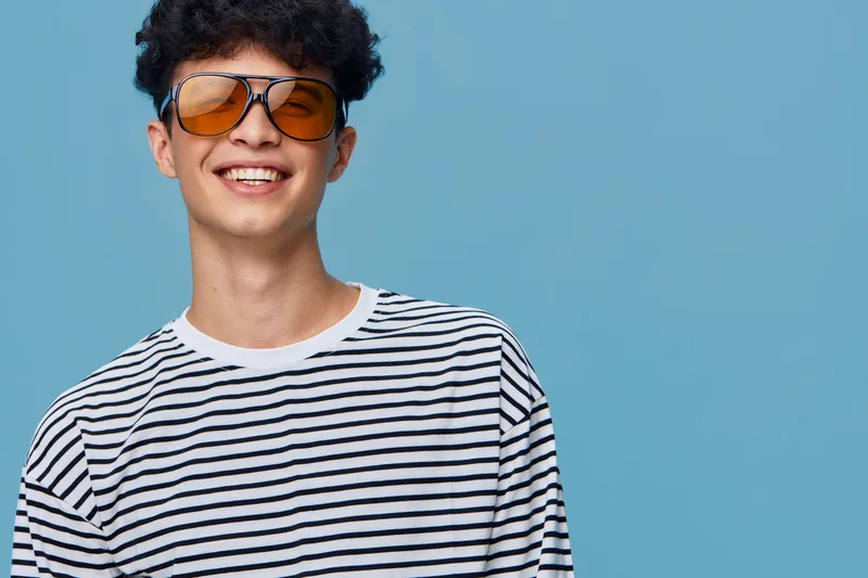 Smiling man wearing orange sunglasses and striped shirt against blue background, expressing happiness and casual summer style.