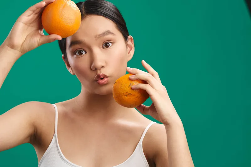 asian woman wearing a white top holds two oranges against a vivid green backdrop, creating a playful pose that conveys energy and a fresh, energetic mood.