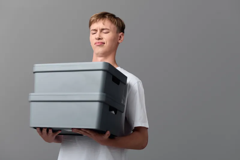 Young delivery enthusiast carries stacked boxes, showing focus and effort, indoor studio setting, clean gray backdrop, minimalism, shipping workflow, organized packaging, logistics theme