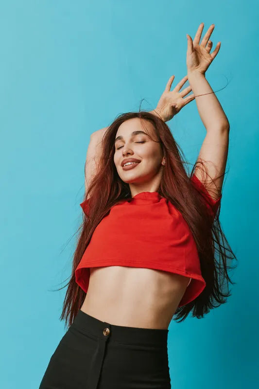 Woman with long hair and closed eyes wearing red crop top and black pants posing with raised arms against blue background. Expression of joy and freedom with casual style.