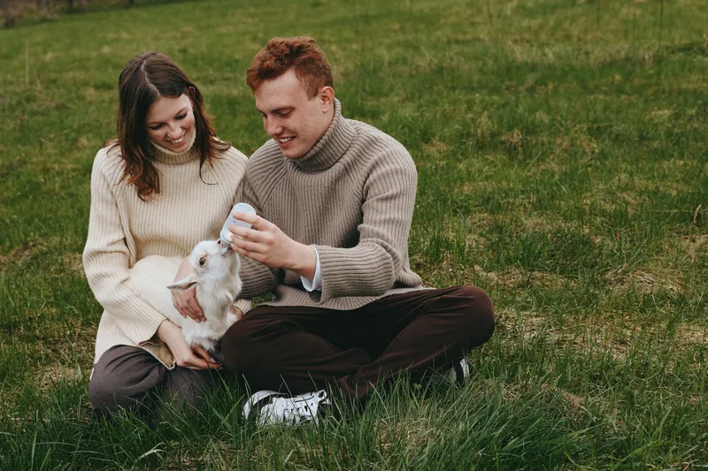 Lifestyle scene with a couple feeding a goat kid outdoors on green grass, sharing affection and smiles in a bright daytime setting, portraying warm family moments outside.