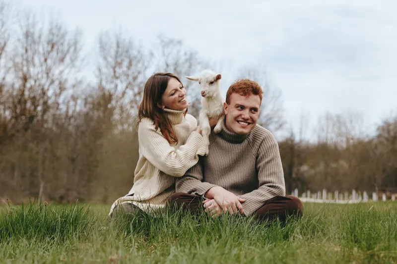 Cheerful couple outdoors with a playful goat kid, shared moment of joy in a sunlit field, cozy sweaters, natural colors, tranquil countryside setting