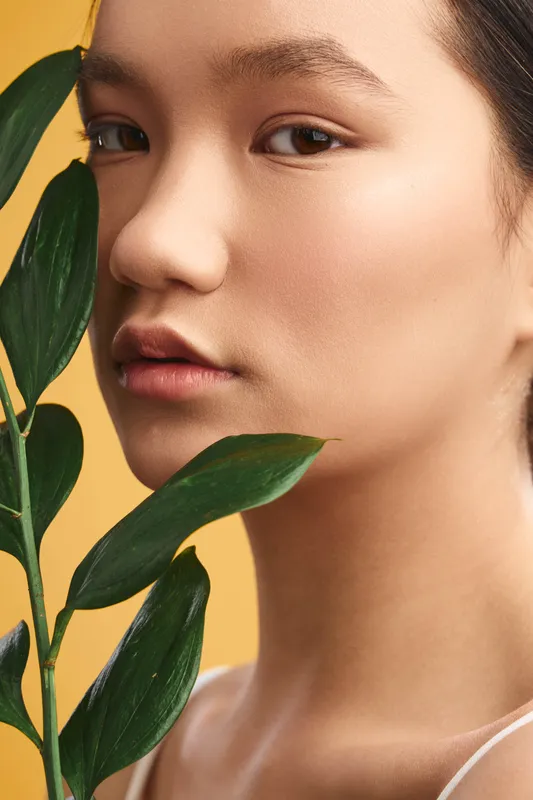 asian woman gazes softly at the camera, framed by leafy stems in a studio setting. warm yellow backdrop enhances her smooth skin and serene mood, conveying calm beauty and natural elegance.