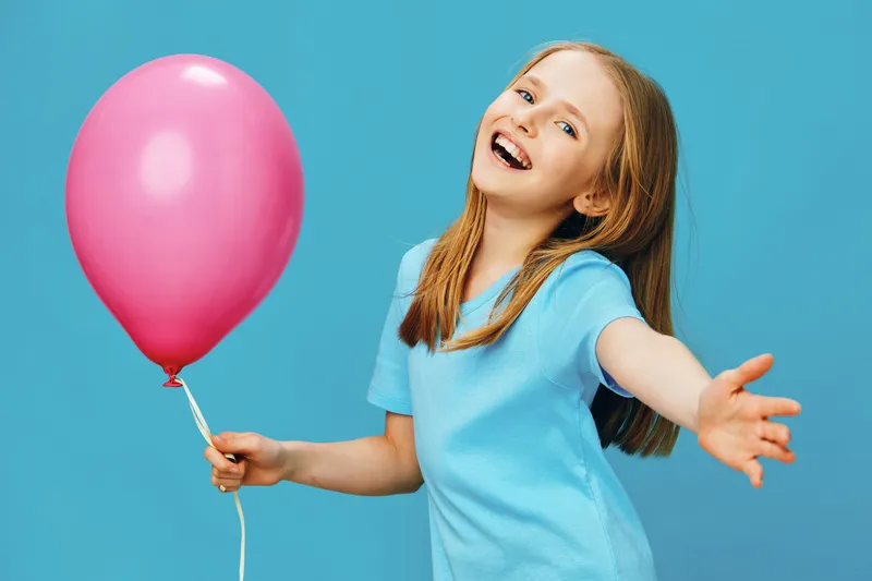 Happy young girl with long hair wearing a blue t shirt holding a pink balloon against a bright blue background, embodying joy and childhood innocence