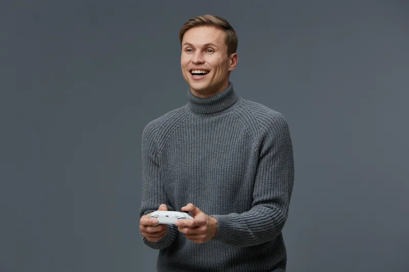 Smiling young caucasian man wearing gray knitted turtleneck playing video game controller isolated on dark gray background digital detox concept