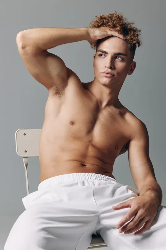 Young man with curly hair sitting shirtless on chair wearing white pants, posing with hand on forehead and looking away in studio with neutral background and soft lighting.