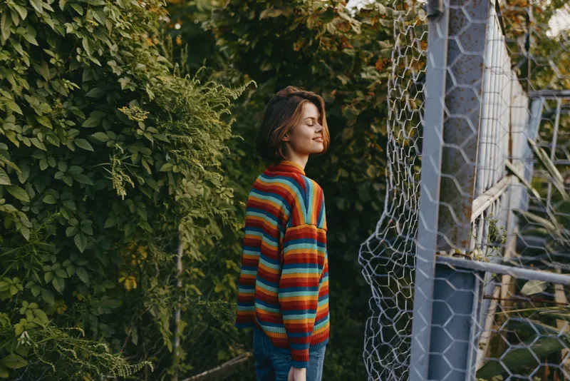 Woman in a colorful striped sweater smiling outdoors by dense greenery and a chainlink fence, young adult in a casual outfit with short brown hair, closed eyes and relaxed posture.