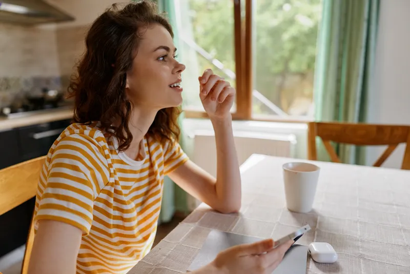 Young woman with wavy brunette hair sits at a sunlit kitchen table using a smartphone and smiling with a mug nearby, creating a warm, relaxed moment in a home setting.