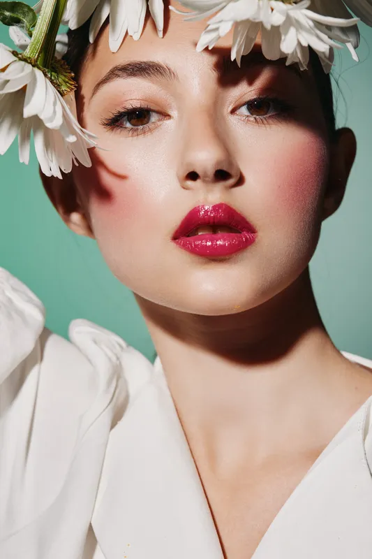 Young woman with radiant skin, red glossy lips, and styled daisies in her hair poses in soft lighting against green background. Creative fashion and beauty concept close-up portrait.