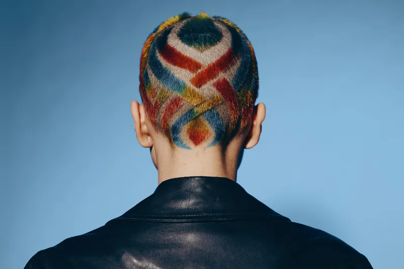 Woman with colorful geometric hair design seen from behind, wearing a black leather jacket against a plain blue background, showcasing bold and creative style.