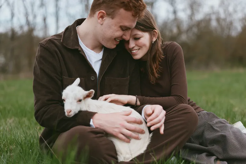 Joyful couple outdoors, tender moment with a goat kid on their lap, warm sunlight, relaxed posture, natural colors, green meadow backdrop, rustic charm, intimate connection, warmth, together.