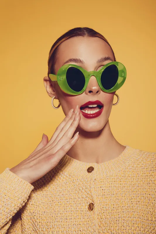 Young woman with green round sunglasses and red lipstick expresses surprise with open mouth and hand near face. She wears yellow knitted sweater on yellow background. Fashion and emotion concept.