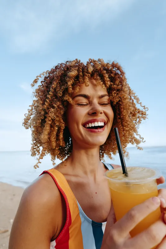 Woman with curly hair smiling on beach, holding drink with straw, summer vacation, healthy lifestyle concept, sunlight, close up portrait, refreshing beverage, ocean background, soft breeze