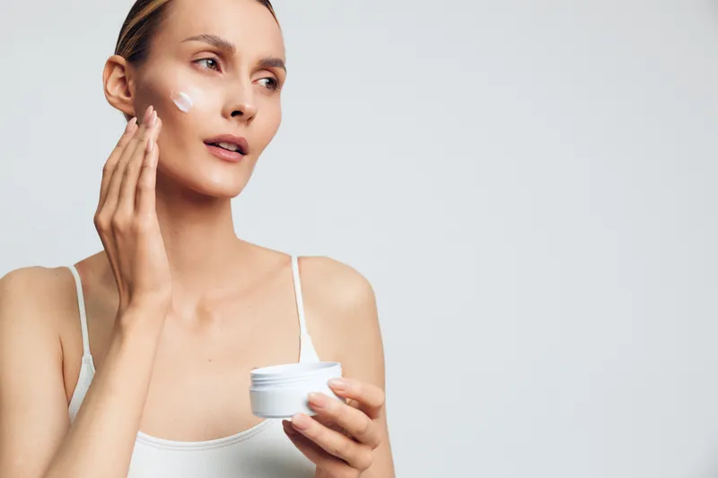 Young woman applies facial cream with fingertips, holding a skincare jar. Bright studio background highlights beauty care routine, selfcare, and healthy skin concept.