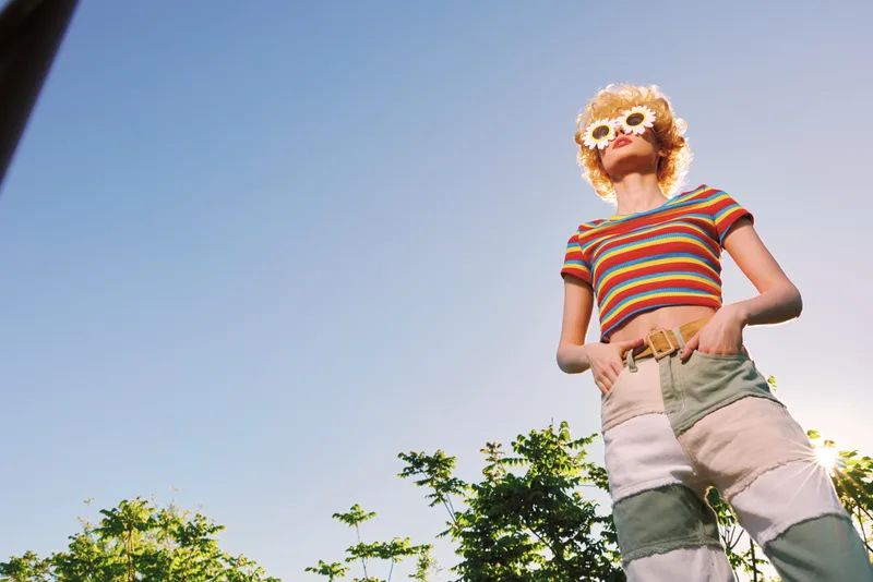 Woman in retro fashion poses outdoors under a clear sky, striped top, patchwork shorts, flower mask sunglasses, vibrant colors and a playful summer lifestyle vibe