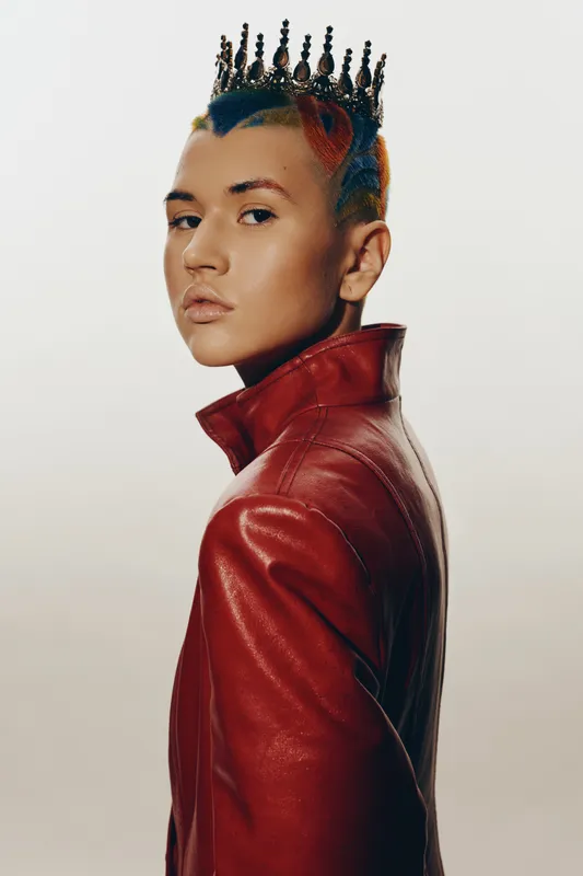 Stylish woman wearing a red leather jacket and a unique black crown with colorful hair, posing with a serious expression against a plain light background.