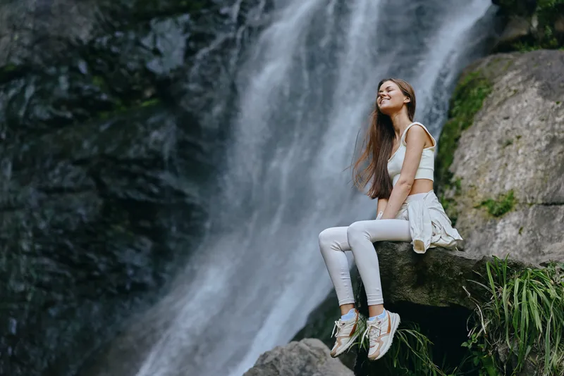 woman sitting near waterfall with casual clothes, enjoying nature, relaxation, peaceful outdoor scene, fresh air, greenery surrounding