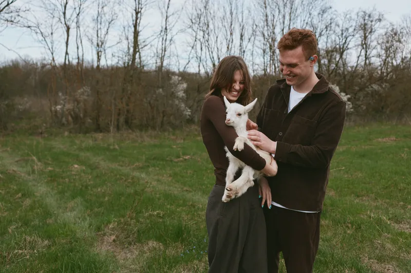 Couple outdoors with a smiling goat kid in a grassy field, warm light, relaxed pose, candid moment, bucolic setting, natural colors, gentle breeze, heartfelt connection