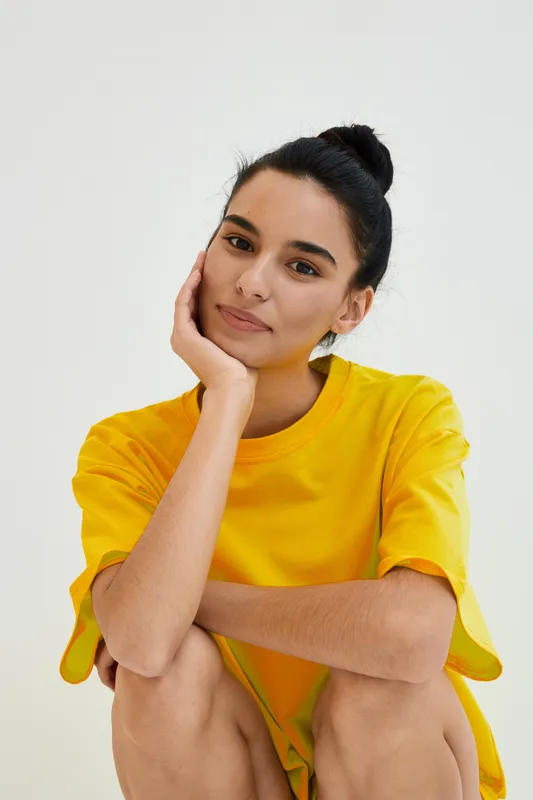 young woman with dark hair in yellow shirt sitting thoughtfully with hand on face against plain white background, casual style, natural expression