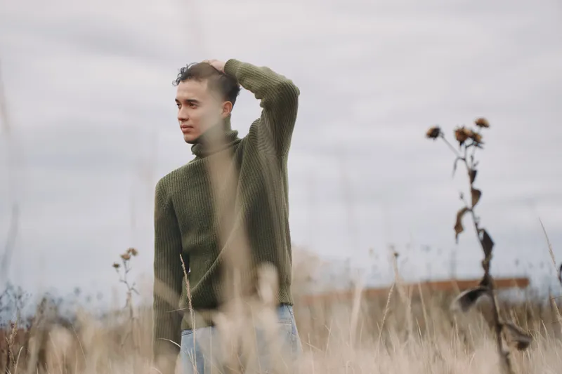 Young man in a green knit sweater standing in a dry field, contemplative gaze conveying authenticity and reliability in a documentary style, natural light, candid outdoor portrait with muted tones.