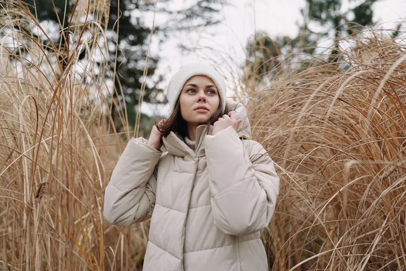 Winter portrait of a woman in a beige padded jacket standing among tall dry grasses, calm expression, soft natural light, outdoor scene conveying solitude and seasonal mood.