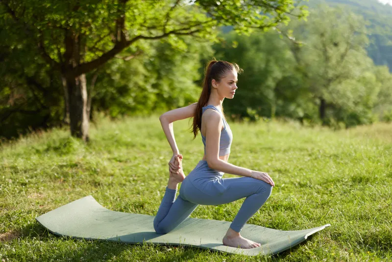 Young woman practicing yoga in a serene green park, wearing a light gray outfit, showcasing flexibility and concentration while meditating outdoors in nature