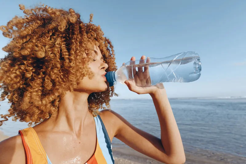 Young woman with curly hair drinks water bottle on a sunny beach, a calm sea and blue sky behind her, conveying hydration and outdoor activity.