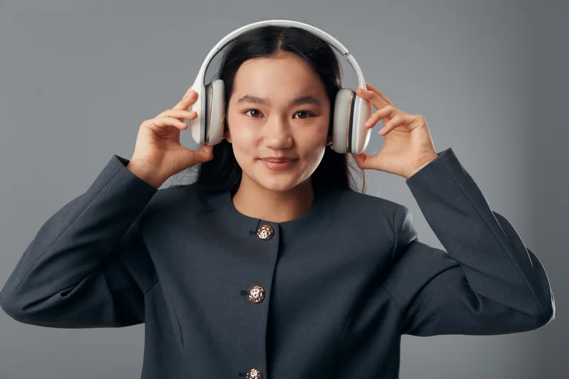 Young woman wearing headphones and smiling against gray background, posing with hands holding device for music listening, entertainment and technology use indoors.