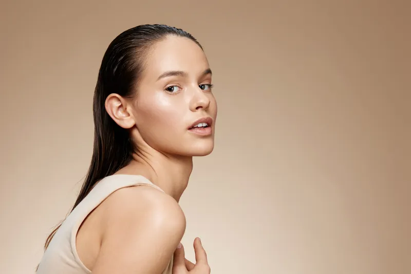 Beautiful young woman with wet hair, glowing skin, and natural makeup, posing against a neutral beige background, embodying confidence and beauty