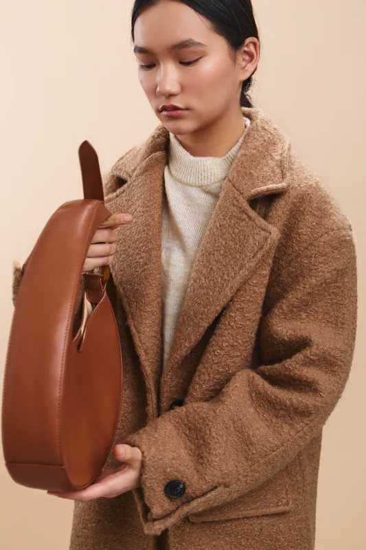 asian woman wearing a textured wool coat holds a brown handbag in a neutral studio setting, looking down with a calm smile, presenting a chic, everyday fashion moment.