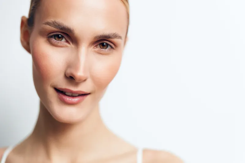 Young woman with clear skin and light makeup smiles gently in a close-up portrait against a white background conveying natural beauty and confidence.