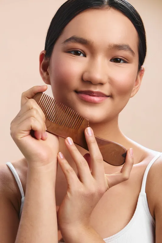 Asian woman smiling softly while holding a wooden comb near her hair in a bright studio, presenting a calm, confident grooming moment filled with warmth and everyday elegance.