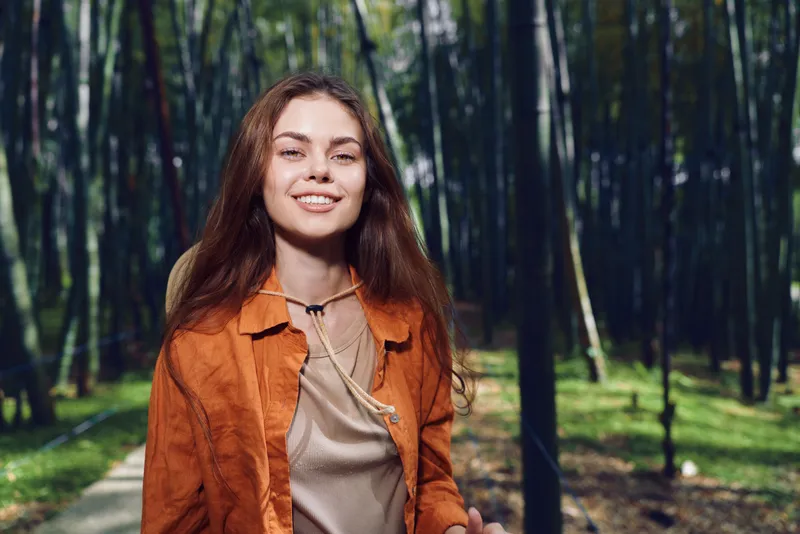 Woman portrait smiling in forest nature, casual fashion jacket and necklace, happy young adult walking on path, sunlight through trees, relaxed outdoor lifestyle portrait.