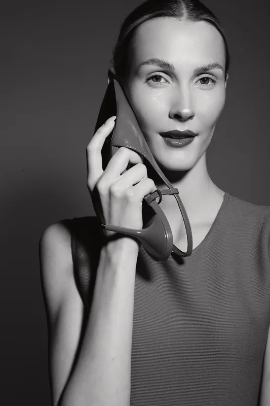 Caucasian woman with light skin holds shoe near face in studio portrait. Concept of fashion, style, and elegance with monochrome lighting.