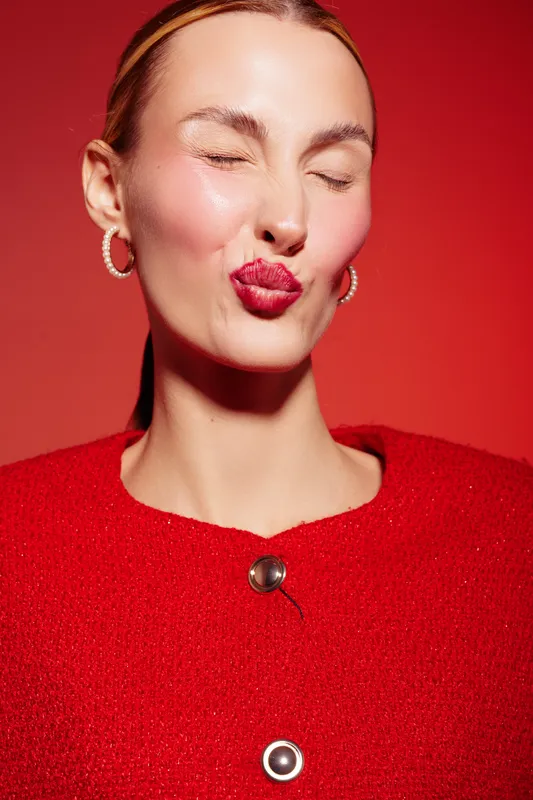 Young woman in red sweater with closed eyes and puckered lips sends a kiss on red background. Concept of affection, love, and playful emotions in studio portrait.