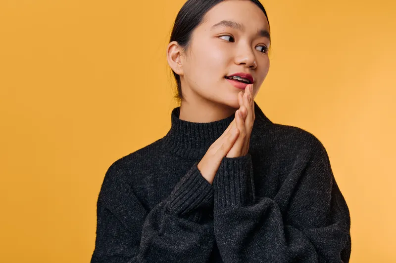 Young woman with dark hair wearing black sweater looking sideways with hands near face on yellow background. Portrait of thoughtful female model with natural makeup and casual style.