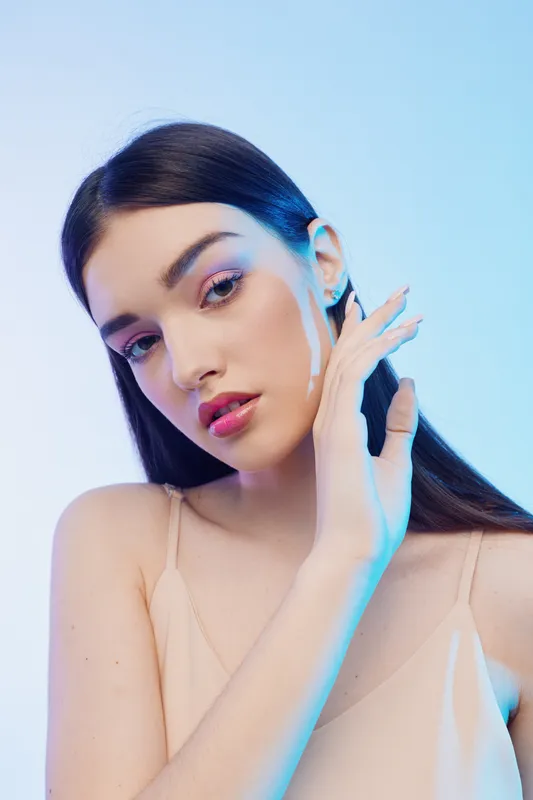 Young woman with long dark hair poses with hand near face in soft beige top. Studio shot with blue background. Concept of beauty, fashion, and elegance.